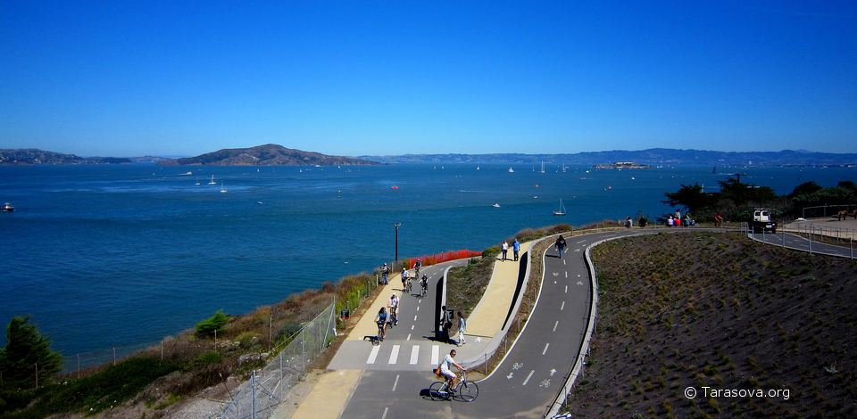 Велосипедисты поднимаются по дорожке к мосту и, затем, пересекая его попадают на противоположный берег залива